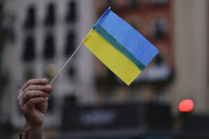 A hand holds up a small Ukrainian flag, which has a blue top half and a yellow bottom half