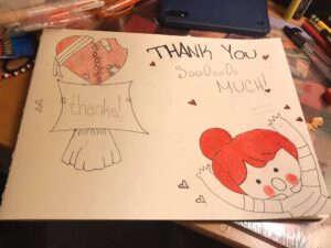 A white piece of paper has a children's thank you note drawn on it, with hearts, and a girl with red hair reaching out her arms. The words say "thank you so so so so much!" 