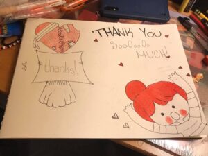 A white piece of paper has a children's thank you note drawn on it, with hearts, and a girl with red hair reaching out her arms. The words say "thank you so so so so much!"