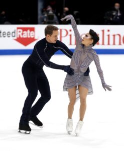 Chock is in a pale purple sparkly costume, with metallic shiny make-up. Bates is in a near-black one-piece outfit, holding onto her waist as if about to spin her around. They are on the ice during the olympics. Chock reaches up into the air with one hand.