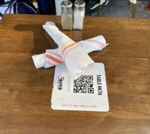 A QR code is partially covered by crossed silverware wrapped in dining napkins on a wood dining table. a salt and pepper shaker are visible behind them. 