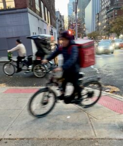 A motion-blurred delivery worker rides his e-bike onto the sidewalk. He wears a blue puffy jacket and carries a red delivery bag on his back.