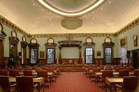 A large room with green wallpaper and a highly decorated cream ceiling. Red Carpet, with red velvet chairs lead up to a dark wood dais, where council members often sit for hearings.