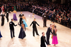 Dancers stand on a brightly lit dance floor in costume, waiting for the music to start. Men are in dark suit-like attire, and women are in long dresses. An audience sits watching, rows deep around the edge of the floor.