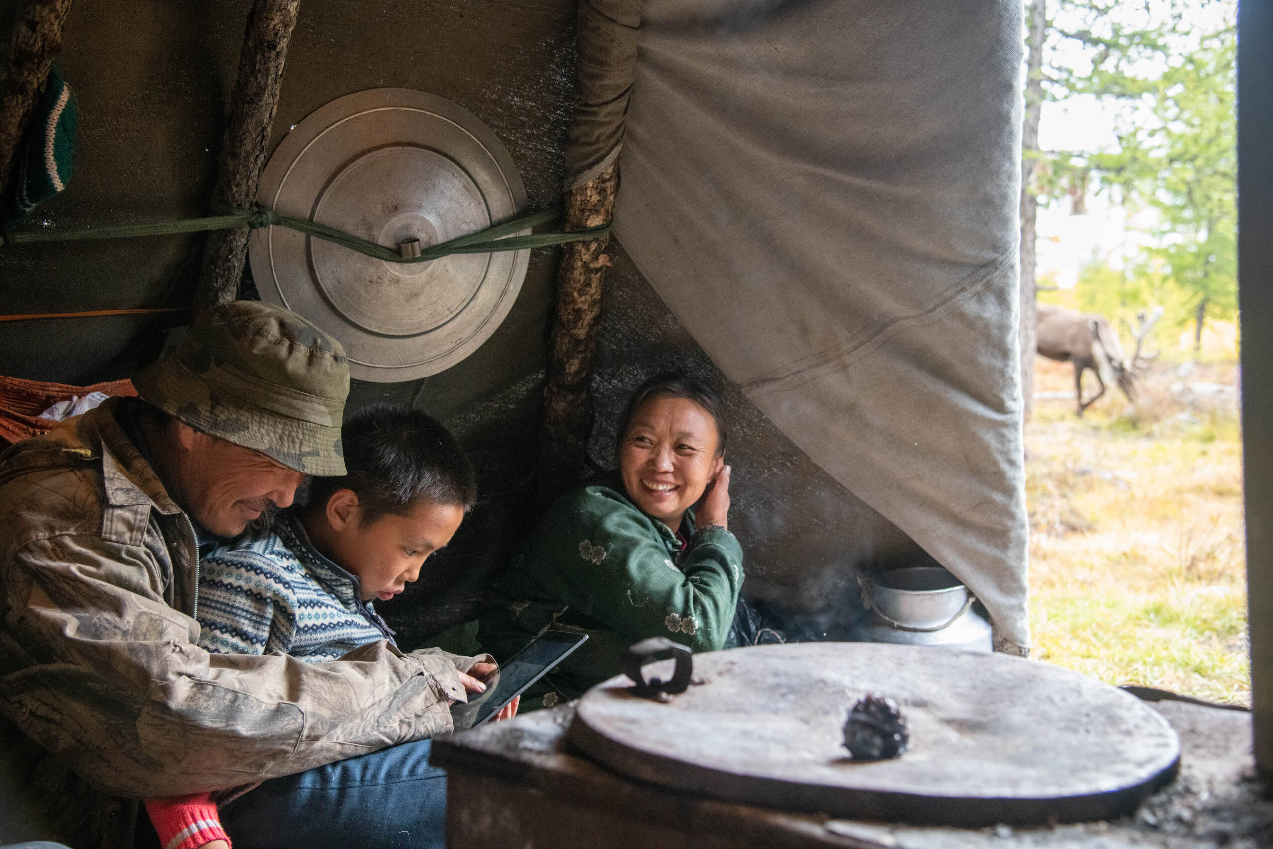 A man holds his son in the foreground as they swipe through images on a tablet together. A woman laughs beside them looking to the left of the frame. He is wearing camo and a bucket hat, and she is wearing a green robe. They are in a tent and there is a stove in the right foreground, and outside the tent flap, some of a reindeer is visible. 