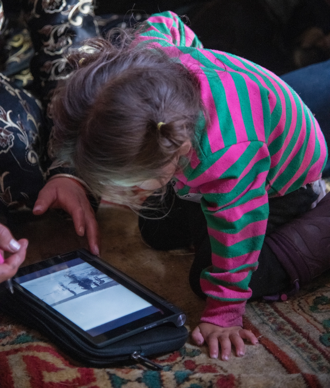 A little girl in pink and green crouches over a tablet.