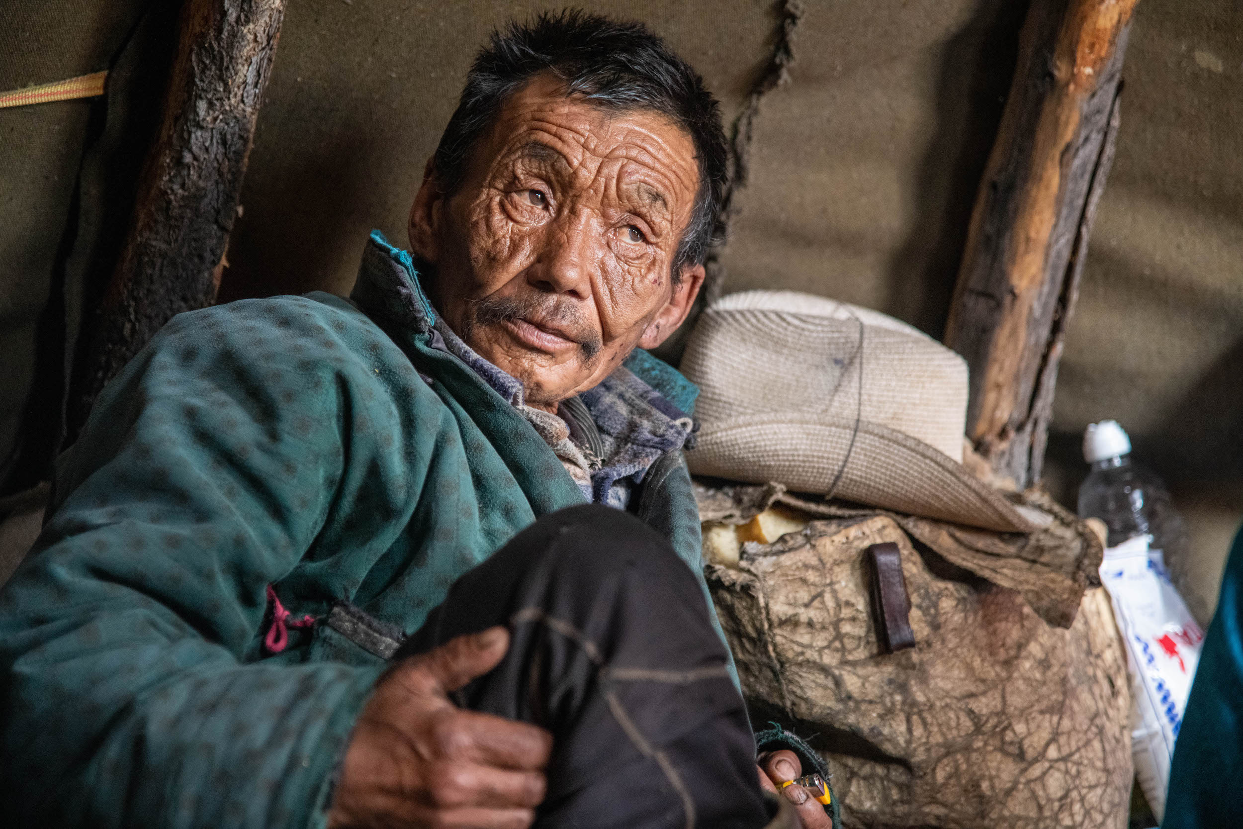 An older man in a green deel or tunic sits inside a tent staring to the right of the frame. There is a cowboy hat and saddle bag behind him.