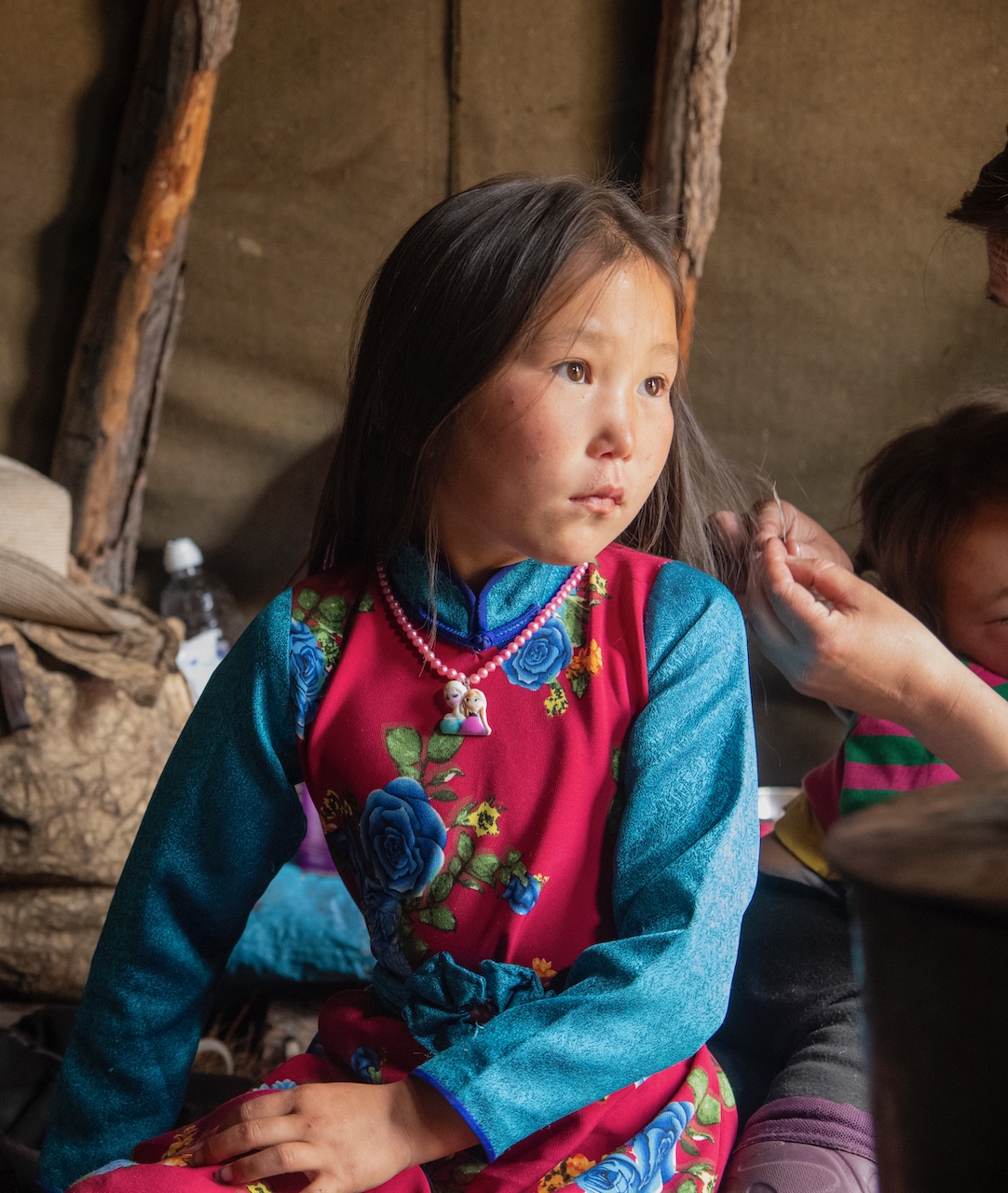 A girl in a colorful, floral tunic of blue and red sits looking to the right of the frame while a woman out of the frame braids her hair.