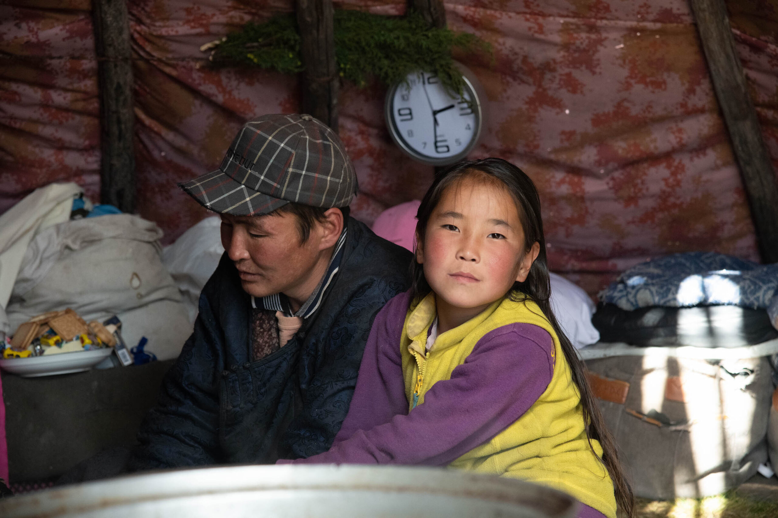 A girl in a yellow vest and a purple sweater looks and the camera while she sits near her father, Galaa. They are in a tent, and he is wearing a baseball cap.