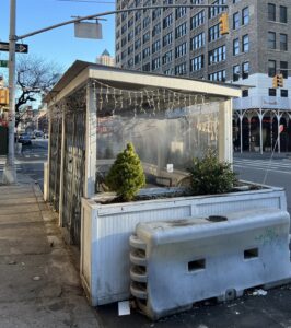 A dirty white dining shed sits on the corner of a Midtown block. The sky is blue and the shines on the shed which has gates locked to prevent homeless from entering. Some evergreen plants line the edge of the shed, and sting lights are attached around the corrugated iron roof.