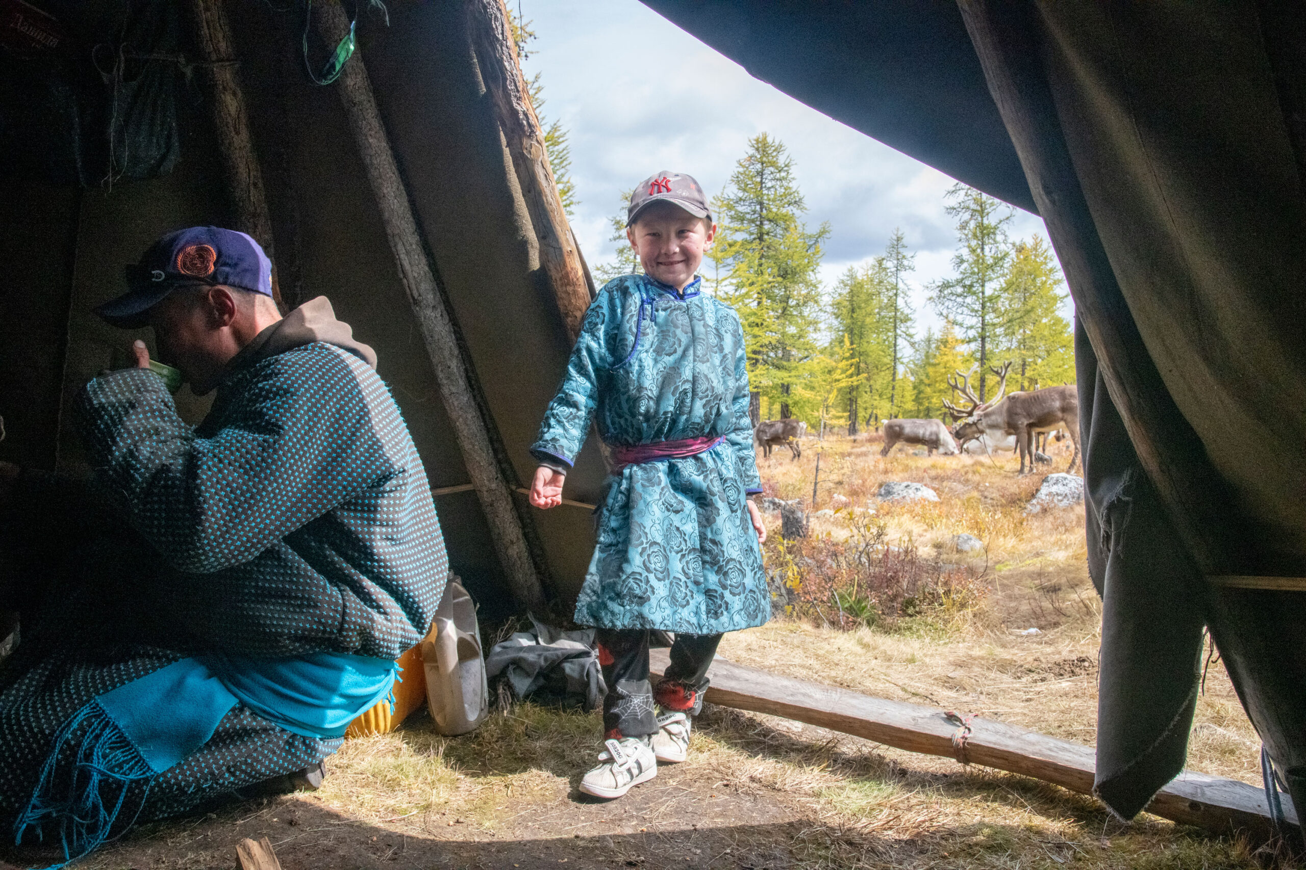 A boy in a blue cloak stands just inside the entrance to a teepee, in the background are trees in the distance, and a man sits drinking from a cup just inside the teepee