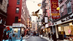 A street in Manhattan's Chinatown, featuring decorated shop windows, awnings, advertisements, and shoppers.