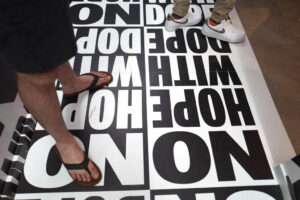 This photo shows a visitors walk through gallery space at the Weedmaps Museum of Weed in Los Angeles. The photo shows two pairs of legs standing on a floor that is covered in a black and white graphic that reads "No Hope With Dope."