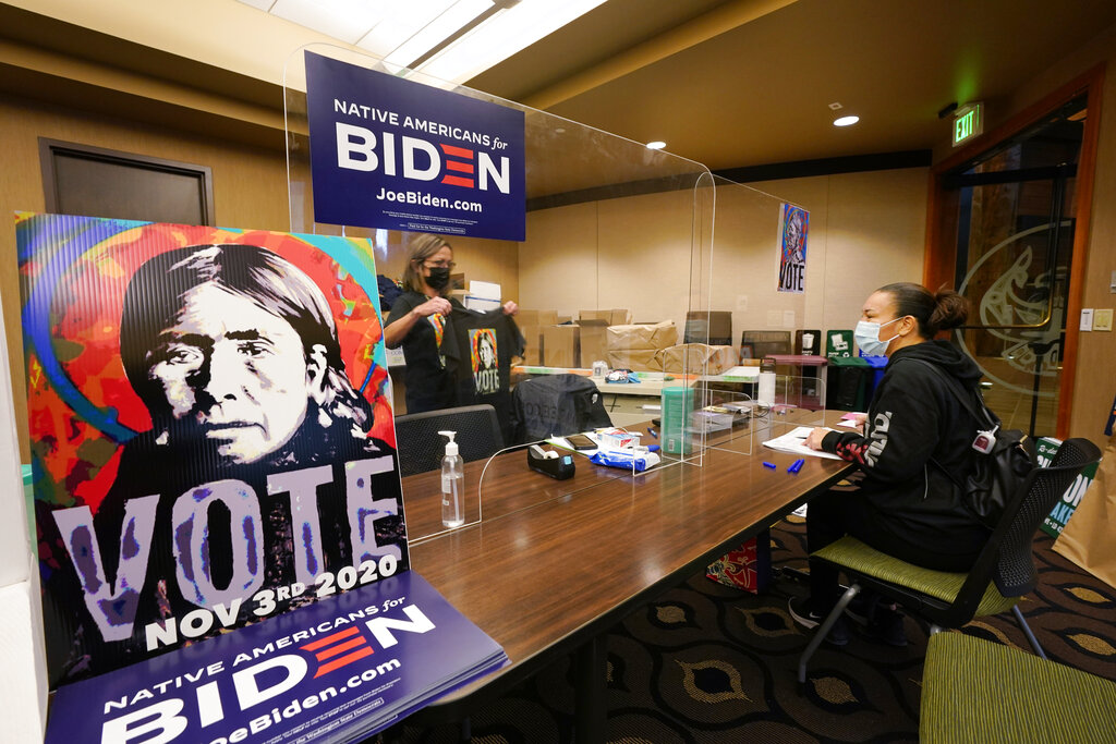 Lummi Tribal member Patsy Wilson is assisted by Lummi Native Vote Team 2020 volunteer Kelli Jefferson in voting. A plastic barrier separates the women. There are "Native Americans for Biden" posters surrounding women.