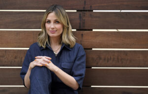 Author Jessica Knoll poses for a portrait. She is sitting against a wooden fence. She is waring navy coveralls.