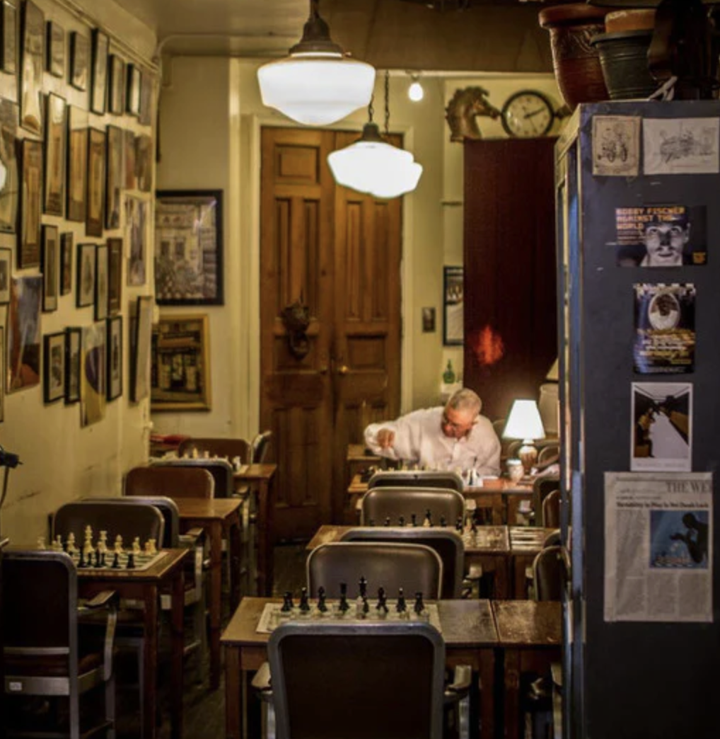 A lone man sets up a chess board on the far end of a room with six chess tables.