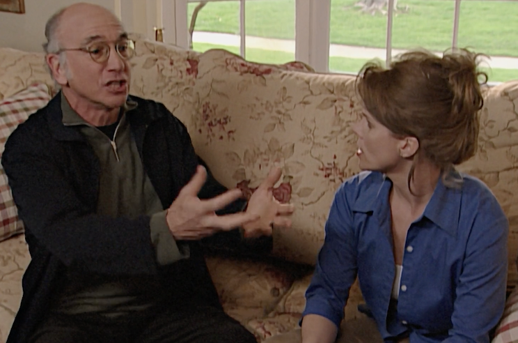 A man sits on a couch, arguing with an off-screen character, his hands thrust forward for emphasis. His wife, sitting next to him, looks on incredulously.