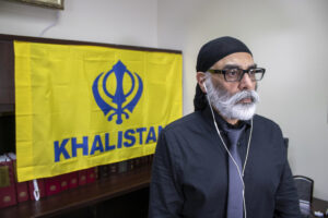 An Indian man sits in front of a yellow flag that has the symbol of a blue Khanda, or double-edged sword, in its center and the word “Khalistan” in blue below it.