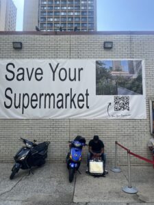 A white banner is fixed to the brick wall of a grocery store. The banner reads “Save Your Supermarket,” and includes a picture of a supermarket as well as a QR code.
