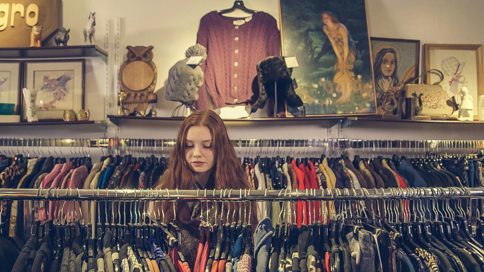 A woman stands in between two racks of colorful shirts in a thrift store.