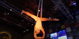 An ariel artist hangs upside down with their legs in a straddle during their performance.