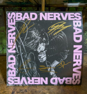 A large pink text circles around a square album cover with the name "Bad Nerves." The gold-colored signatures from the band members are written across the album cover. The lead singer sings into a microphone in black and white. The album cover sits atop of a granite table.