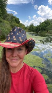 Morris smiles in a photo taken by herself. The South San Gabriel River is behind her. She wears a red shirt and a red, white, and blue cowboy hat. The hat has stars and stripes like U.S.A. flag. The cloudy blue sky is reflected in parts of the river not covered in algae.