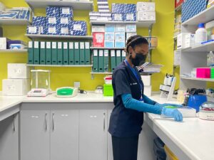 A masked and gloved Yemaachi Biotech Research Associate clears a counter in front of shelves of lab material.