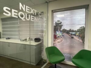 There are two green chairs in front of a window looking out to the streets of Accra, Ghana. A window beside has “Next Gen Sequencing” in decals. 