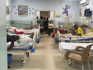 Two rows of hospital beds with outpatients line each side of the children’s ward. People celebrate a child’s birthday at the end of the hallway.