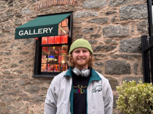 A man dressed with a green and white jacket, black shirt with colored letters, and green hat stands in front of a stone wall. A silver pair of headphones is hung around his neck as he smiles into the camera. A green bush is next to him.