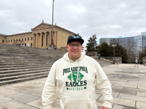 A man wearing a white hooded sweatshirt with the words "Philadelphia Eagles" and "Football" written across it with a black hat, including a green eagle in the front, posing at the Rocky Steps and smiling at the camera. The man is also wearing glasses.