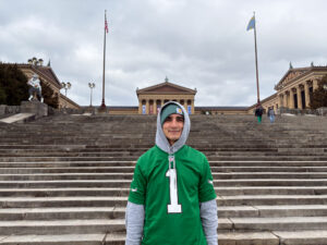 A man wearing a green Eagles jersey over a grey long-sleeved sweatshirt with a green hat is posing at the Rocky Steps and smiling in front of the camera.