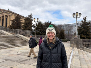 A woman wearing a green and white hat and black jacket is posing at the Rocky Steps and smiling at the camera.