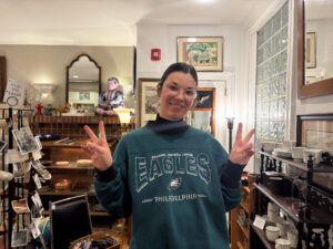 A woman dressed in a green sweater with the words "EAGLES" and "Philadelphia" written across it is posing with two peace signs up and smiling in front of the camera. The woman is also wearing glasses with kitchenware and furniture behind her.