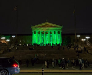 A group of Eagles fans dressed in black, white, and green gear walk up the Rocky Steps at night. The pillars of the Philadelphia Museum of Art are lit up in green against the dark sky.