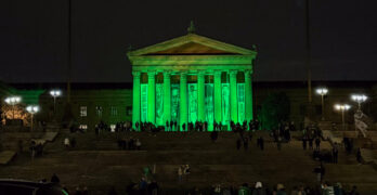 A group of Eagles fans dressed in black, white, and green gear walk up the Rocky Steps at night. The pillars of the Philadelphia Museum of Art are lit up in green against the dark sky.