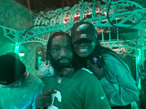 A man dressed in a green jersey at a restaurant smiling at the camera. The entire picture is green. A woman is wrapping her hand around the man's neck and leaning her head against his as she smiles for the camera.